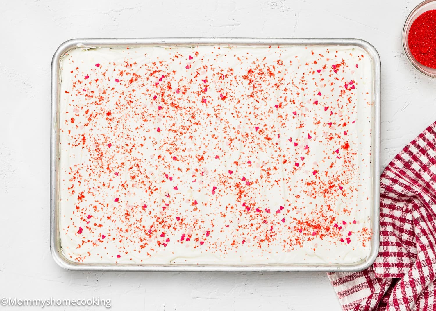Eggless Red Velvet Brownie Poke Cake with a rectangular shape, white frosting, and red sprinkles sits on a baking tray beside a red and white checkered cloth.