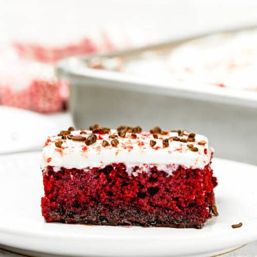 A slice of eggless red velvet cake with white cream frosting and chocolate sprinkles sits invitingly on a white plate.