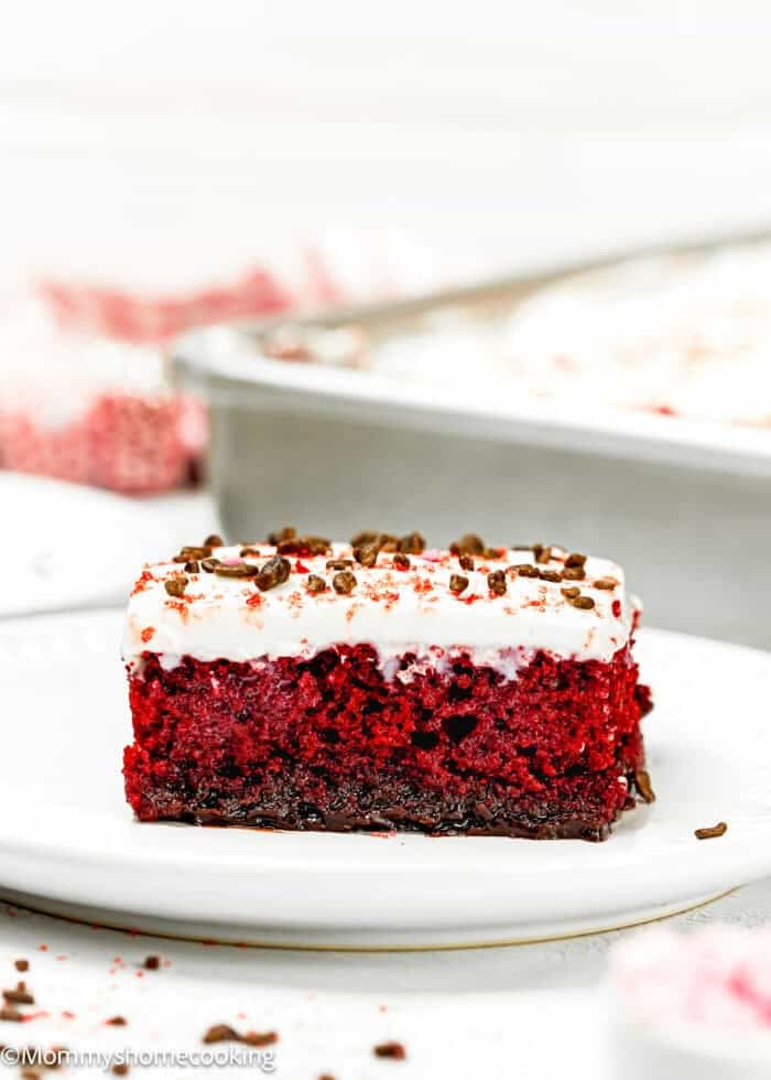 A slice of eggless red velvet cake with white cream frosting and chocolate sprinkles sits invitingly on a white plate.