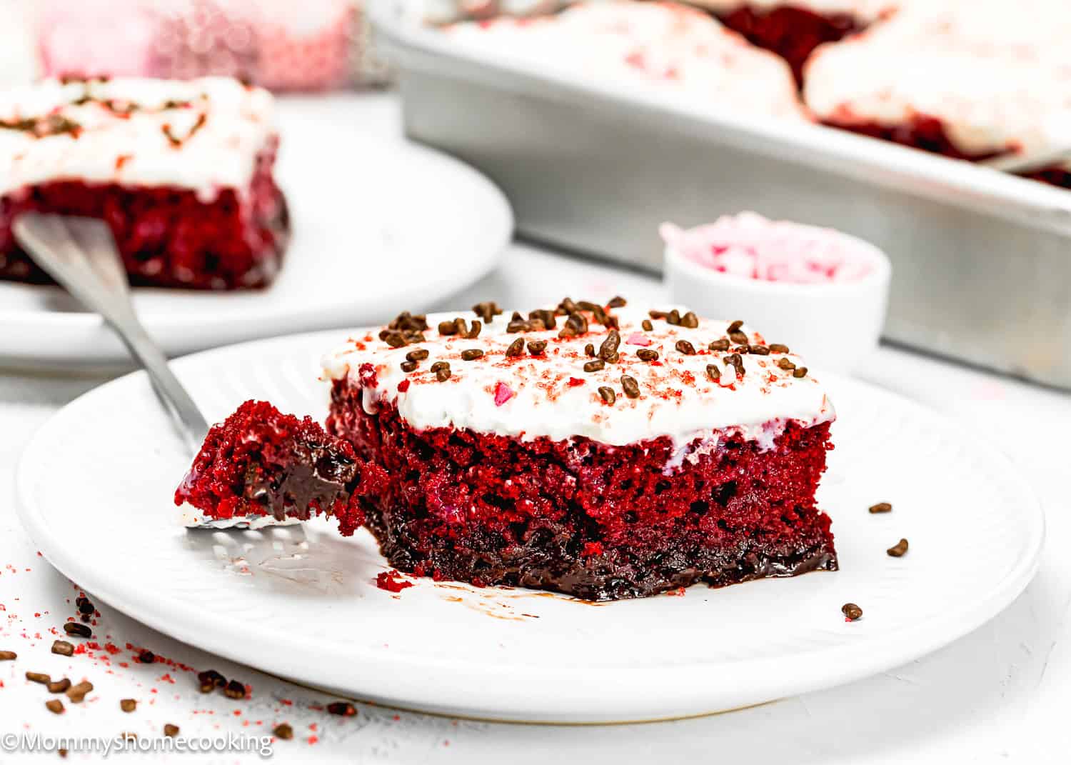 An eggless red velvet brownie poke cake sits elegantly on a white plate, adorned with cream cheese frosting and chocolate sprinkles. Beside it, a fork holds a tempting piece of the decadent dessert, inviting you to indulge in its rich flavor.