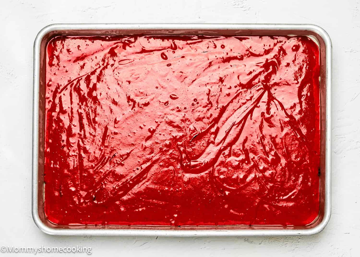 A rectangular baking tray filled with a smooth layer of eggless red velvet poke cake batter.