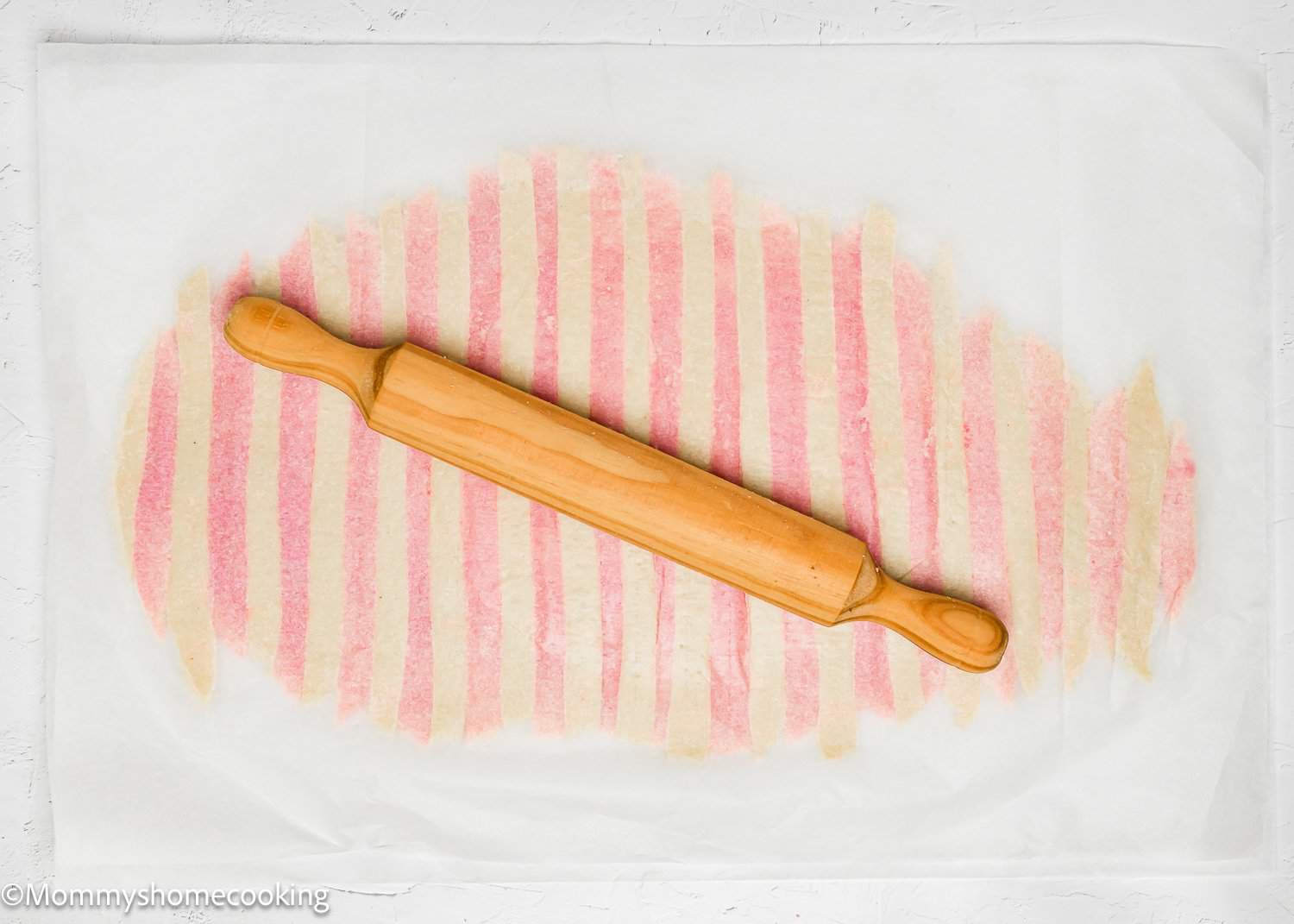A wooden rolling pin rests on parchment paper, ready to craft delightful Eggless Valentine's Striped Sugar Cookies with a charming striped pattern underneath.