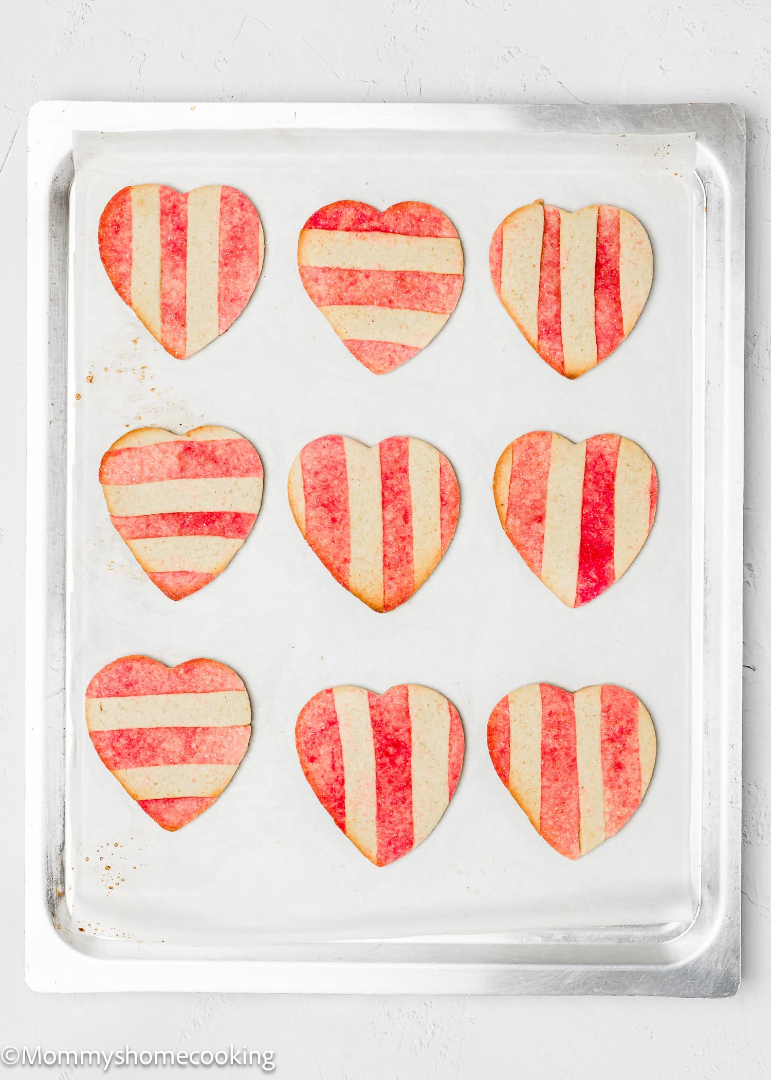Nine eggless Valentine’s sugar cookies with red and white stripes rest charmingly on a baking sheet, ready to delight.