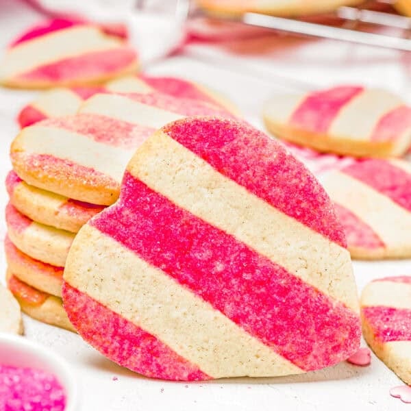 Eggless Valentine's striped sugar cookies, heart-shaped with pink and white diagonal stripes, are stacked in a small pile beside a bowl of pink sugar.
