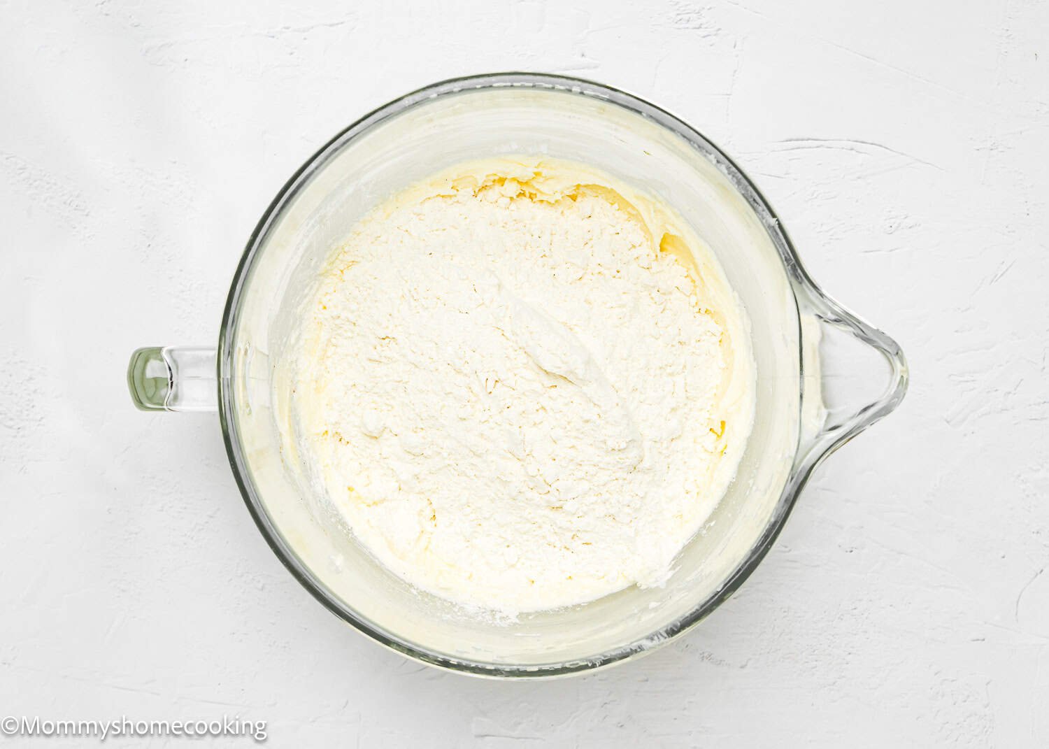 A glass mixing bowl contains white flour resting on top of an eggless creamed butter and sugar mixture, ready to create delightful Valentine's striped sugar cookies against a pristine white background.