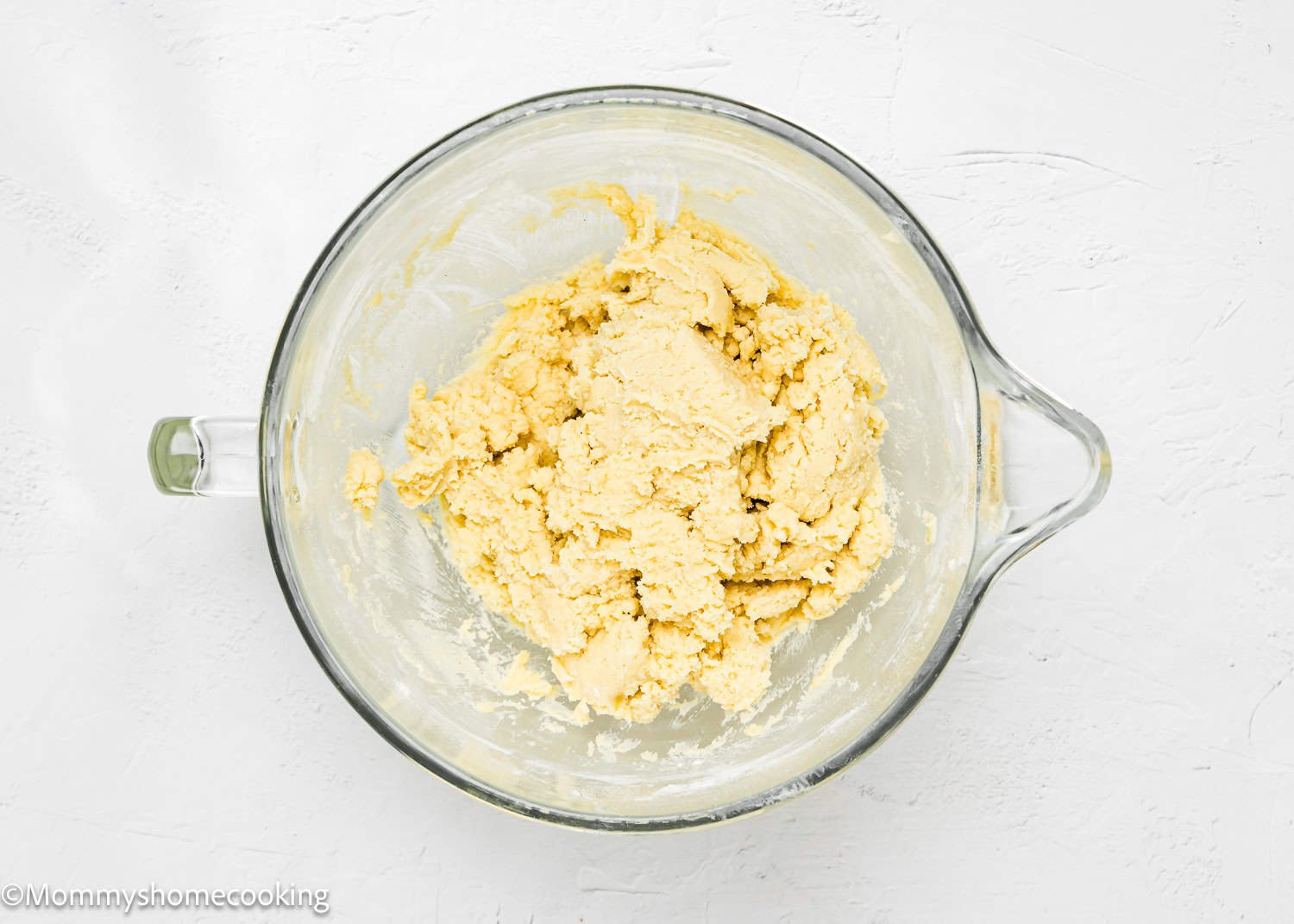 A glass bowl holds a mound of pale yellow eggless cookie dough, perfect for crafting delightful Valentine's striped sugar cookies, on a white textured surface.