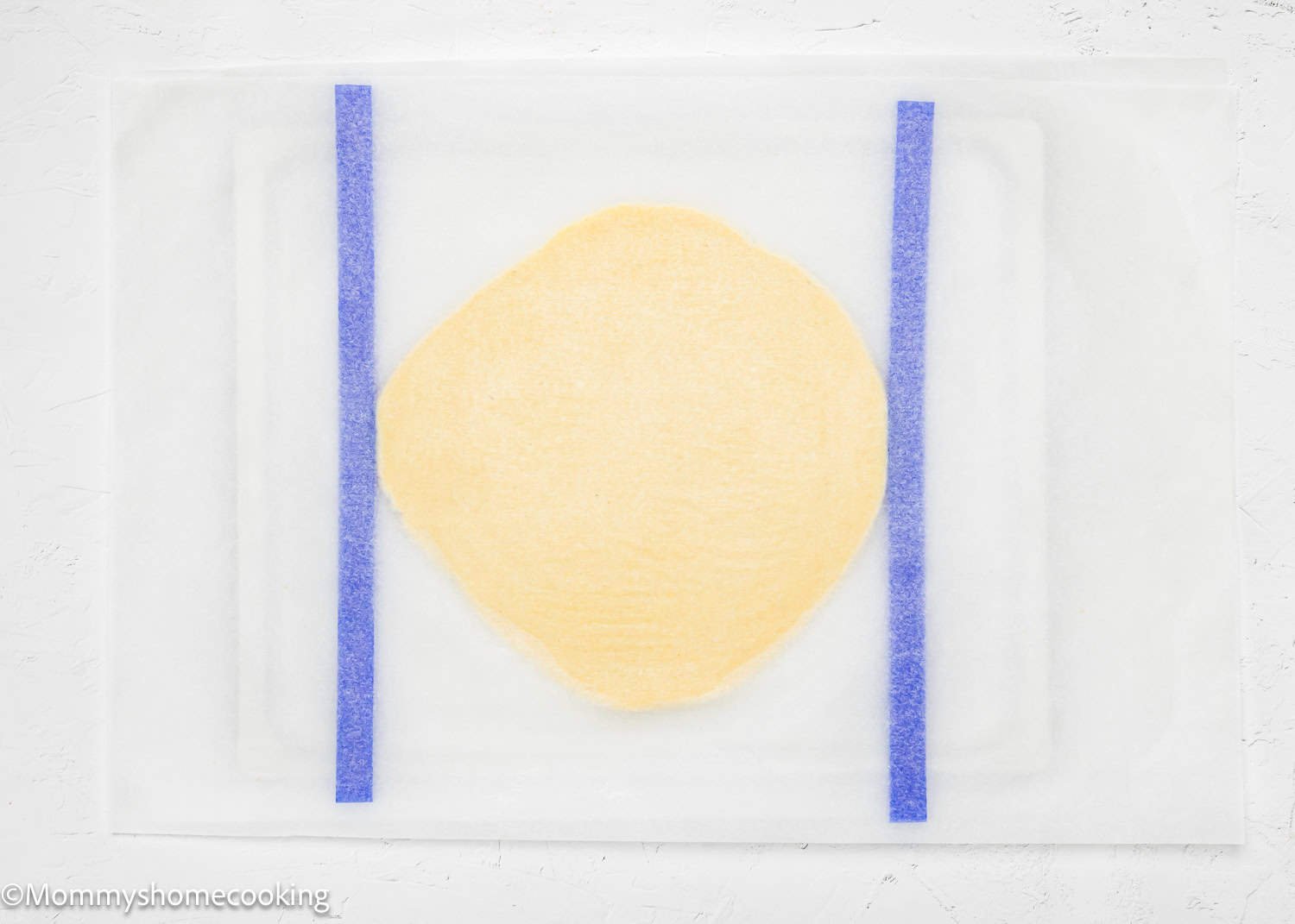 A round piece of yellow dough, perfect for crafting eggless Valentine's striped sugar cookies, is placed between two blue rolling pin guides on a white surface.