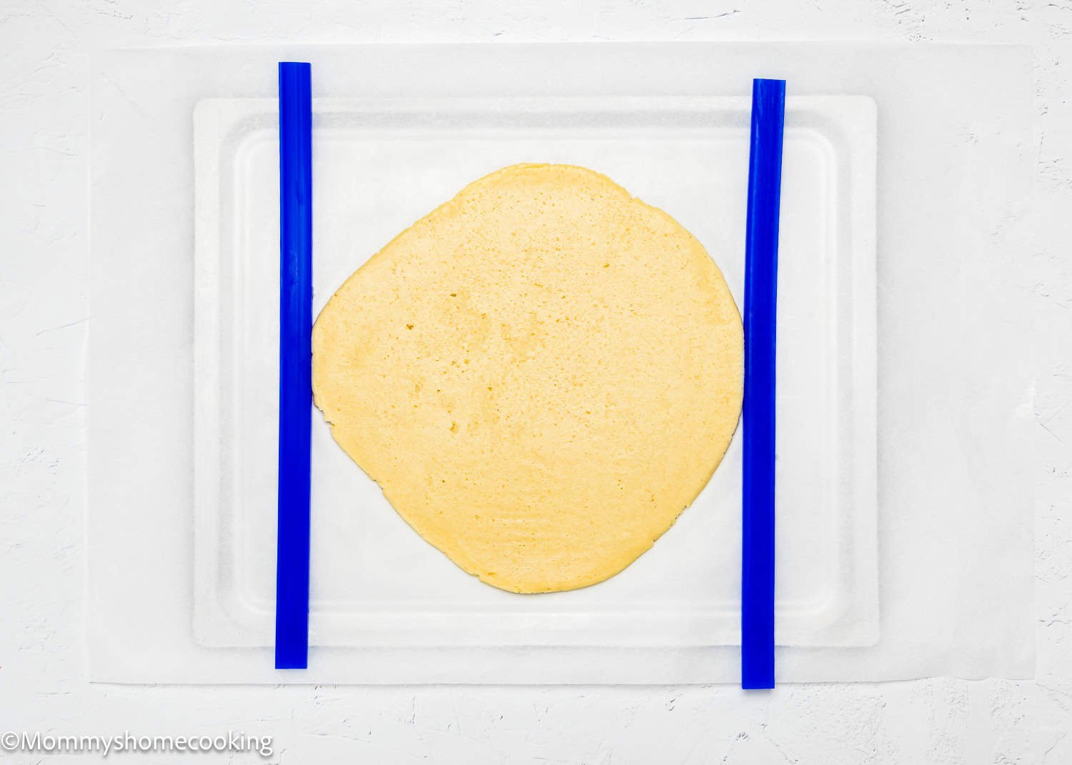 An eggless dough sheet for Valentine's striped sugar cookies is carefully placed between two blue guide bars on a white surface.