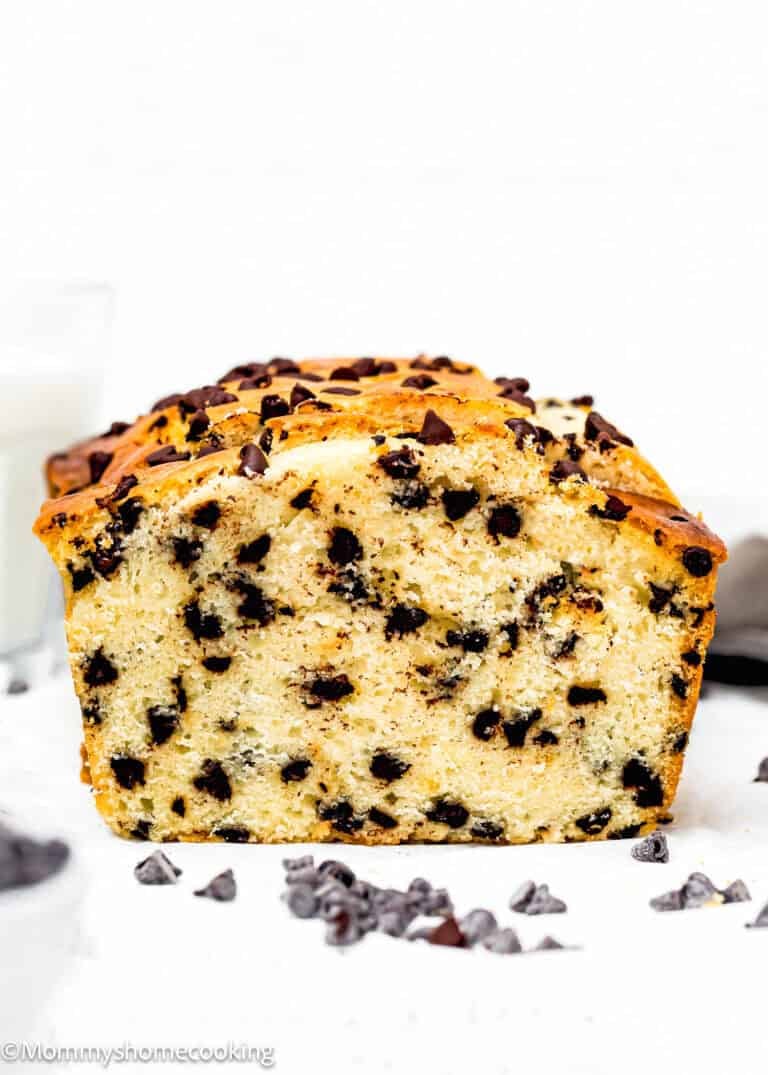 Delight in this fluffy chocolate chip loaf cake on a white surface, with chocolate chips scattered around. This treat is made without eggs or milk, offering a unique twist on the classic dessert everyone loves.