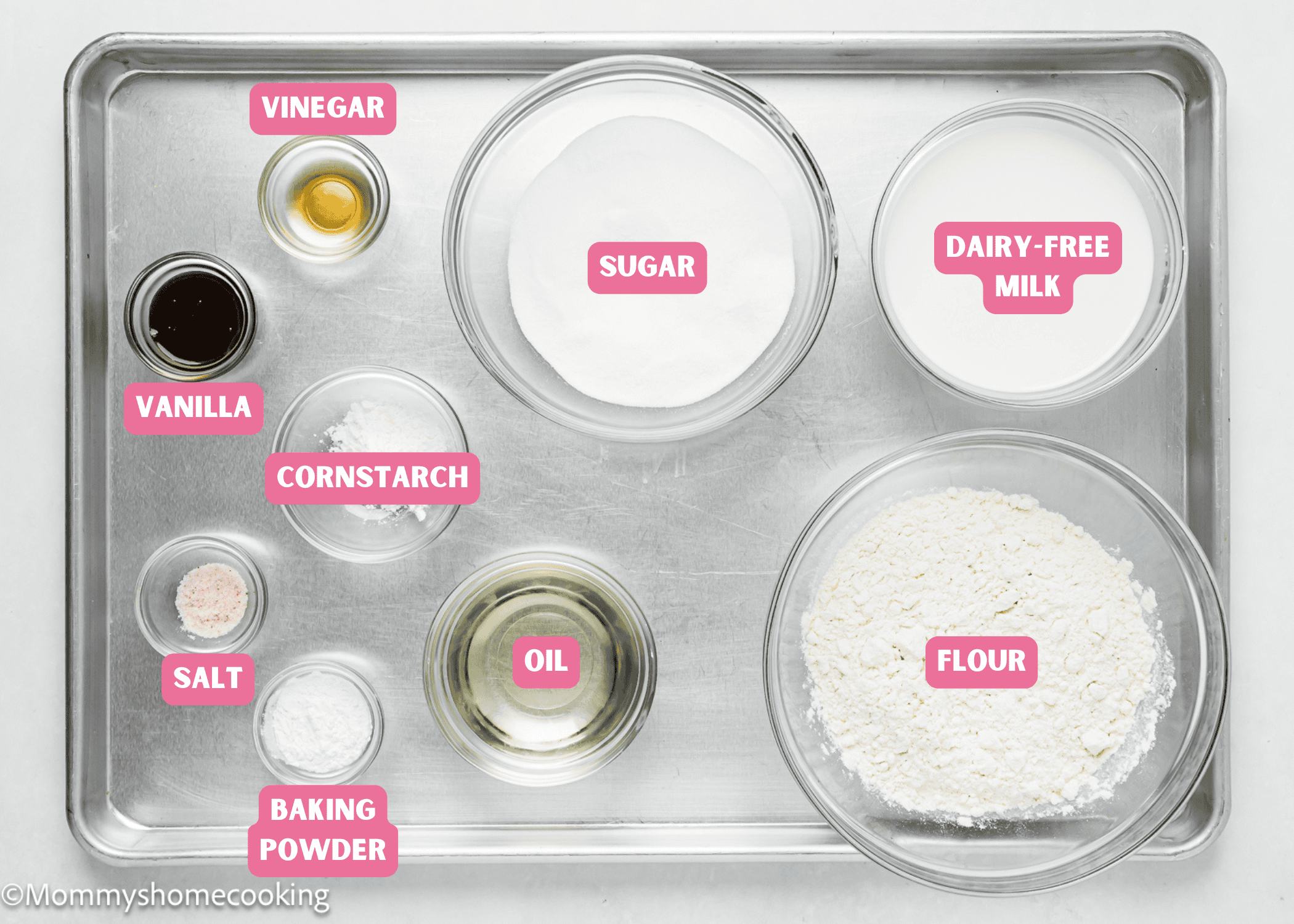 A baking tray with labeled ingredients for an easy, egg-free and dairy-free vanilla sheet cake: vinegar, salt, sugar, cornstarch, oil, baking powder, dairy-free milk, and flour.