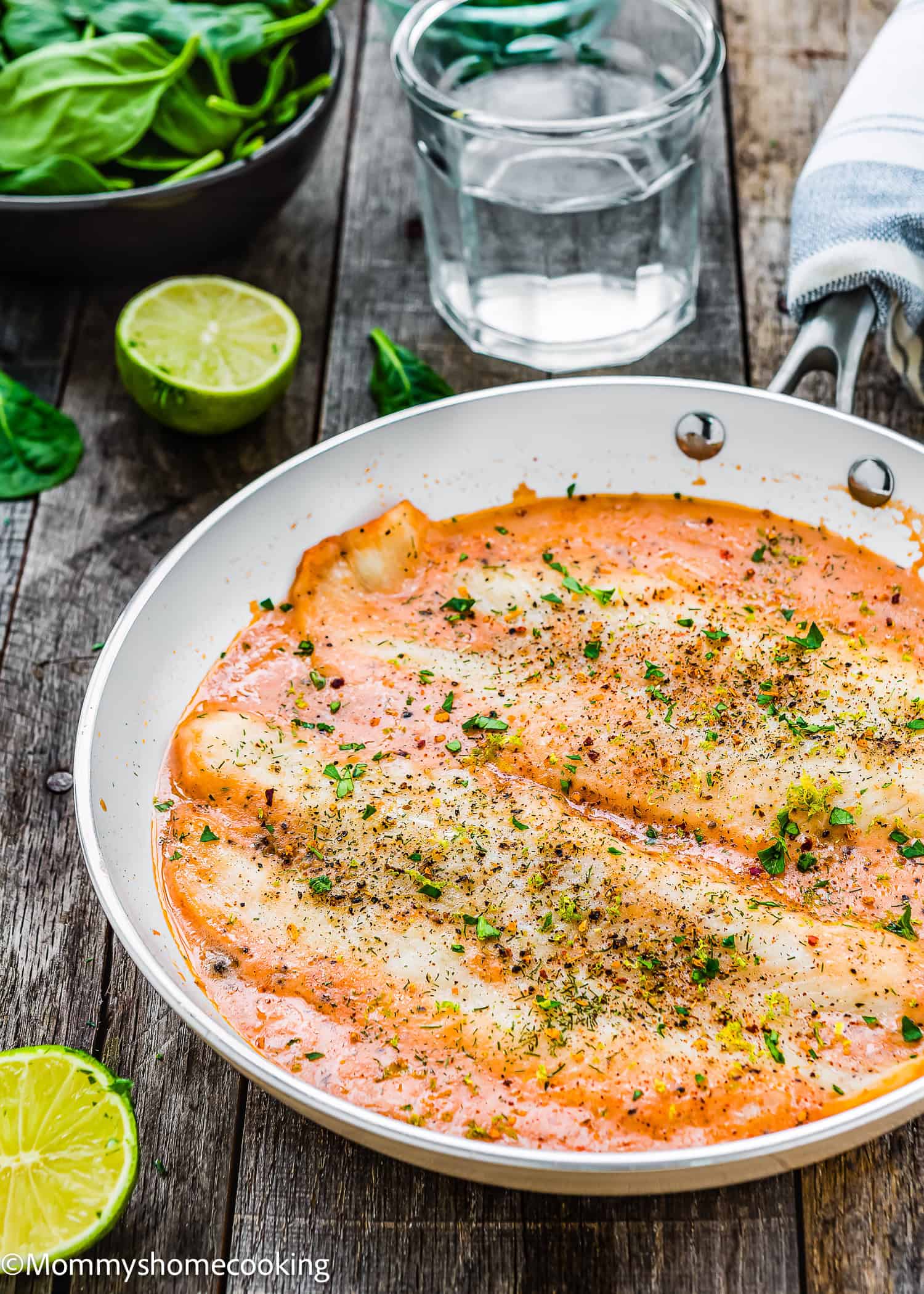 Quick and easy creamy fish fillet casserole with baked fish fillets topped with herb seasoning in a rich tomato sauce, garnished with parsley, served in a pan on a wooden table alongside sliced lime and a refreshing glass of water.