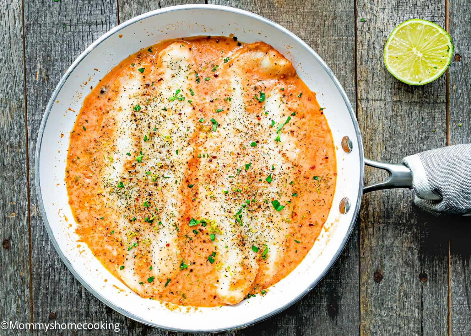 Quick and easy creamy fish fillet casserole in a white skillet on a wooden surface, surrounded by a rich red sauce with herbs and accompanied by a lime half nearby.