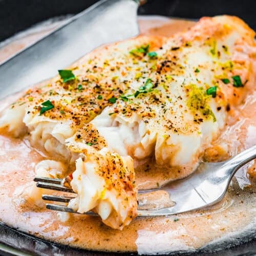 Quick and easy oven-baked fish filet with herbs, served in a creamy sauce on a dark plate. Knife and fork on the side. Lime slice in the background, offering a touch of zest.