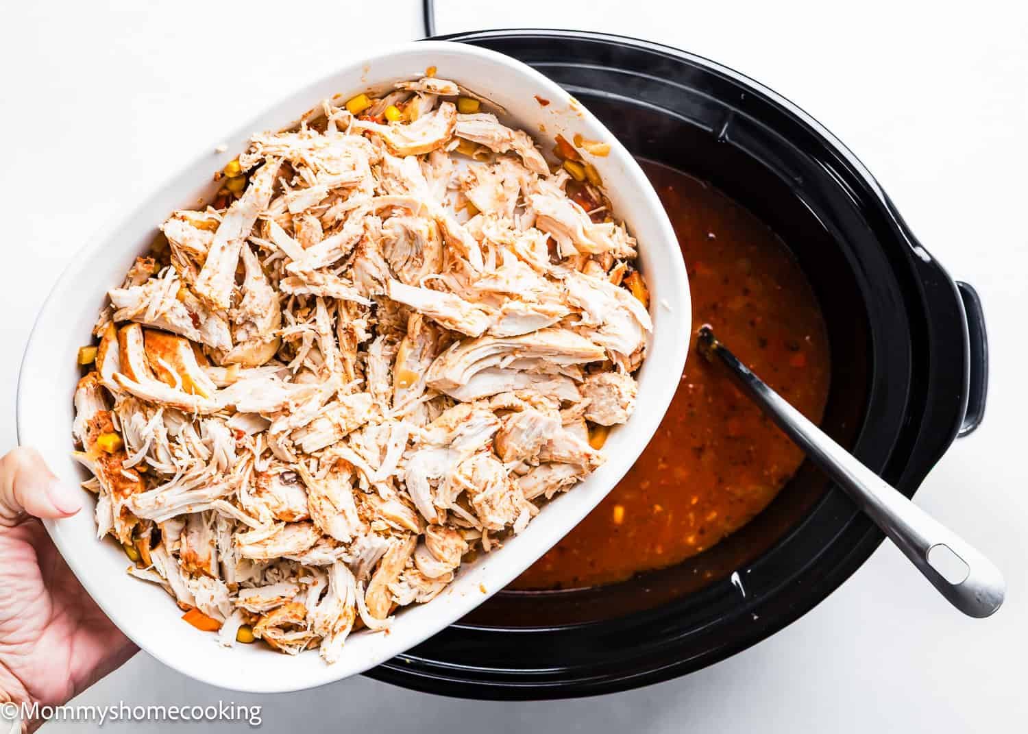 A hand adds shredded chicken from a white dish into a slow cooker filled with rich Tortilla Soup, accompanied by a ladle.