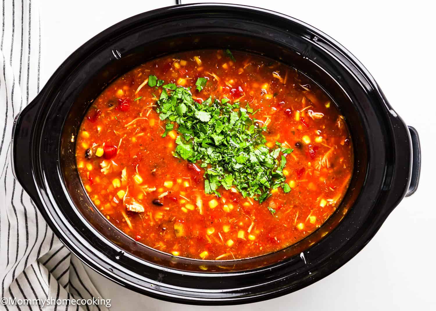 A slow cooker filled with hearty chicken tortilla soup topped with chopped cilantro, containing beans, corn, and chunks of tomato. A striped cloth is placed beside the cooker.