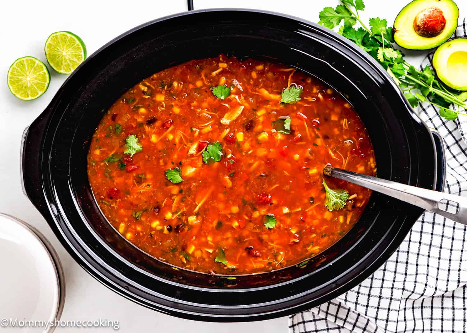 A slow cooker filled with chicken tortilla soup, garnished with cilantro. Nearby are avocado halves, lime slices, and a striped cloth.