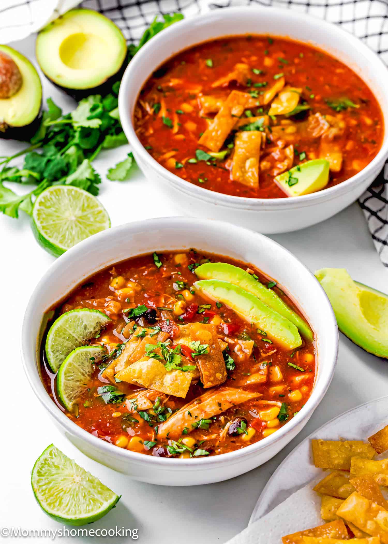 Two bowls of Slow Cooker Chicken Tortilla Soup are garnished with avocado slices, lime wedges, and cilantro, set on a white surface. Nearby are avocado halves and fresh cilantro leaves.