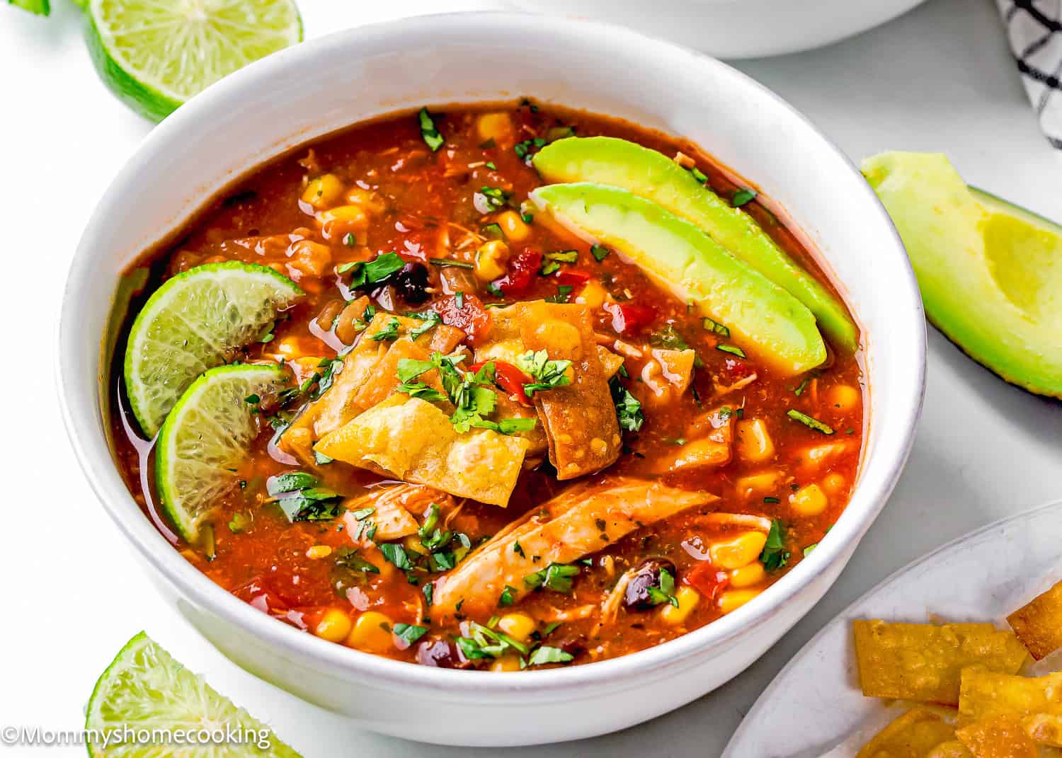 A bowl of slow cooker chicken tortilla soup topped with lime wedges, avocado slices, and crispy tortilla strips, garnished with fresh cilantro.