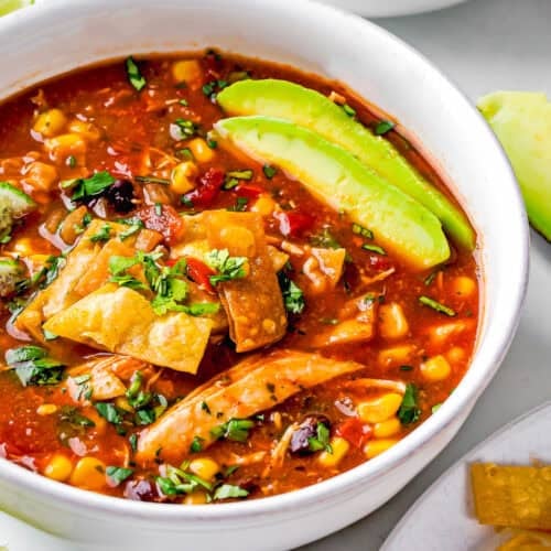 A delicious bowl of slow cooker chicken tortilla soup, topped with fresh avocado slices, crunchy tortilla strips, sweet corn, black beans, and chopped cilantro.
