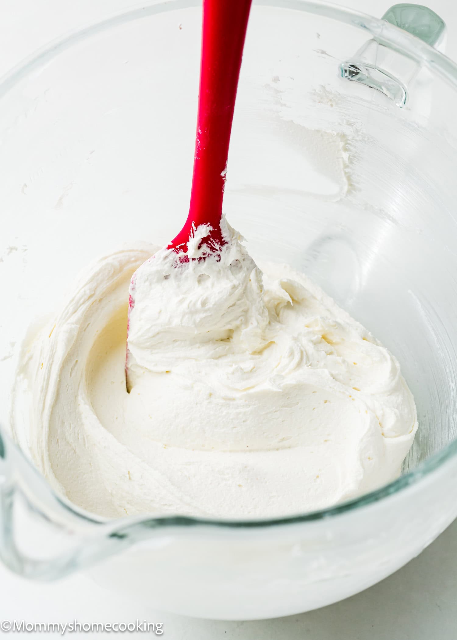 An easy clear mixing bowl holds creamy, vanilla frosting being stirred with a red spatula. Perfect for topping an egg-free and dairy-free vanilla sheet cake.