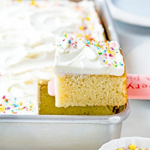 An easy, egg-free and dairy-free vanilla cake with white frosting and colorful sprinkles is being lifted from a baking pan.