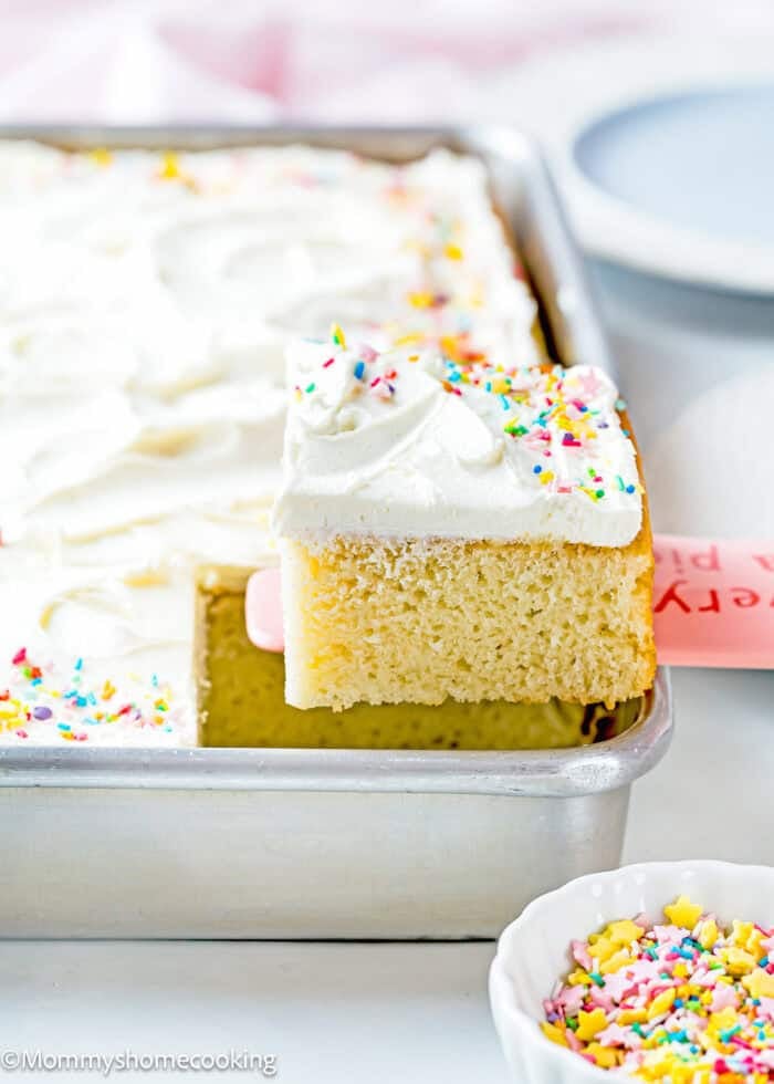 An easy, egg-free and dairy-free vanilla cake with white frosting and colorful sprinkles is being lifted from a baking pan.