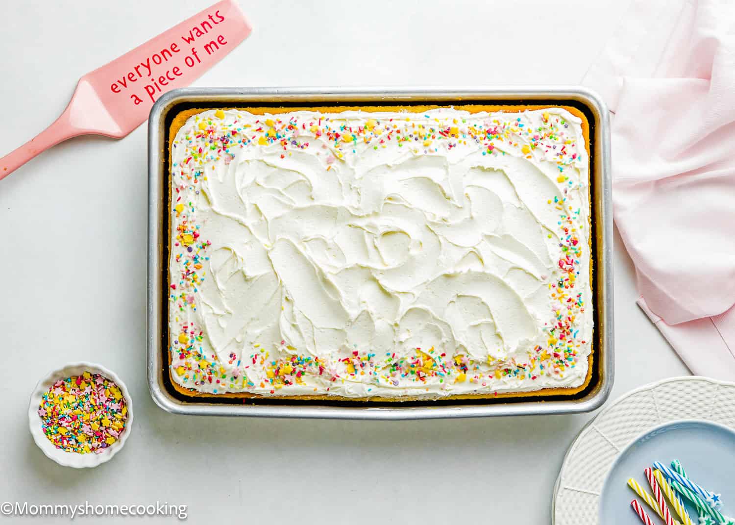 This easy rectangular cake is an egg-free and dairy-free vanilla sheet cake, topped with white frosting and colorful sprinkles on a baking tray. Nearby are a spatula, a small bowl of sprinkles, a stack of plates, and candles.