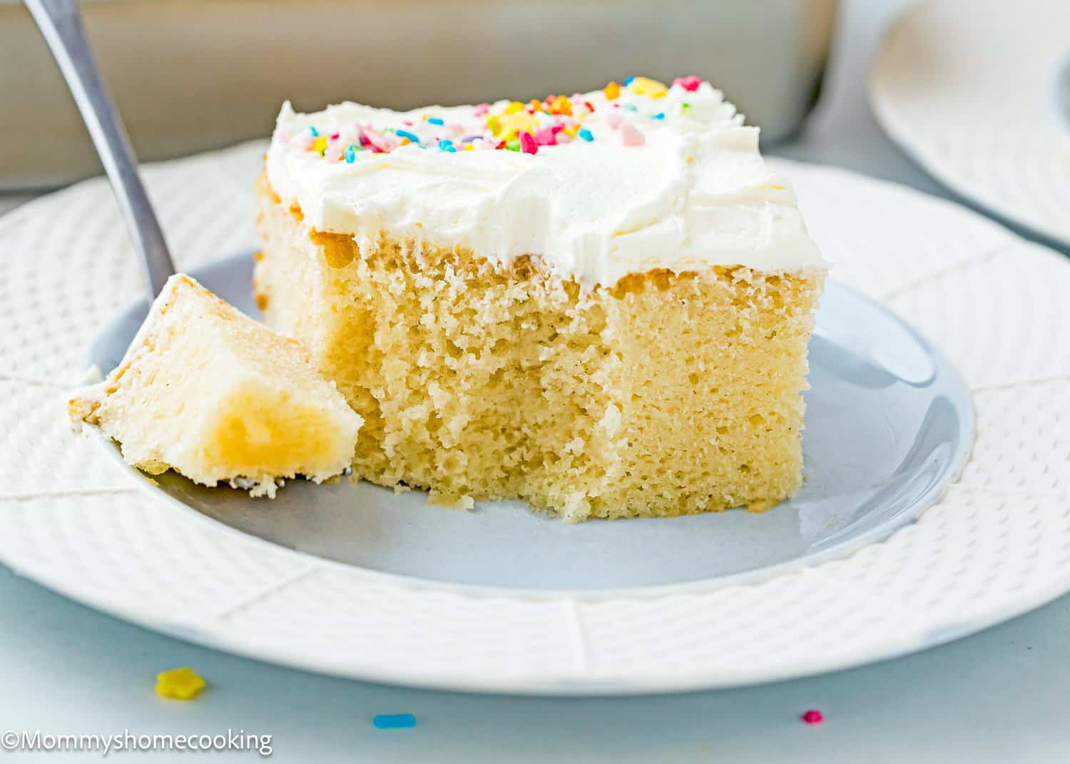 An easy slice of egg-free and dairy-free vanilla cake with white frosting and colorful sprinkles sits invitingly on a plate, with a fork ready to enjoy a piece.