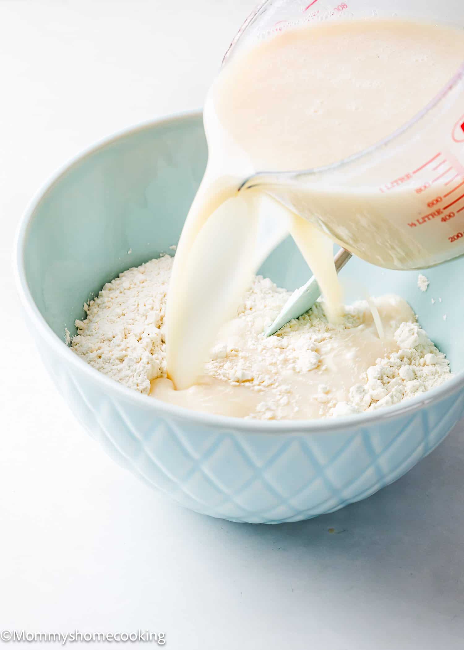Liquid from a measuring cup is poured into a bowl with a flour mixture, perfect for preparing an easy, egg-free and dairy-free vanilla sheet cake.