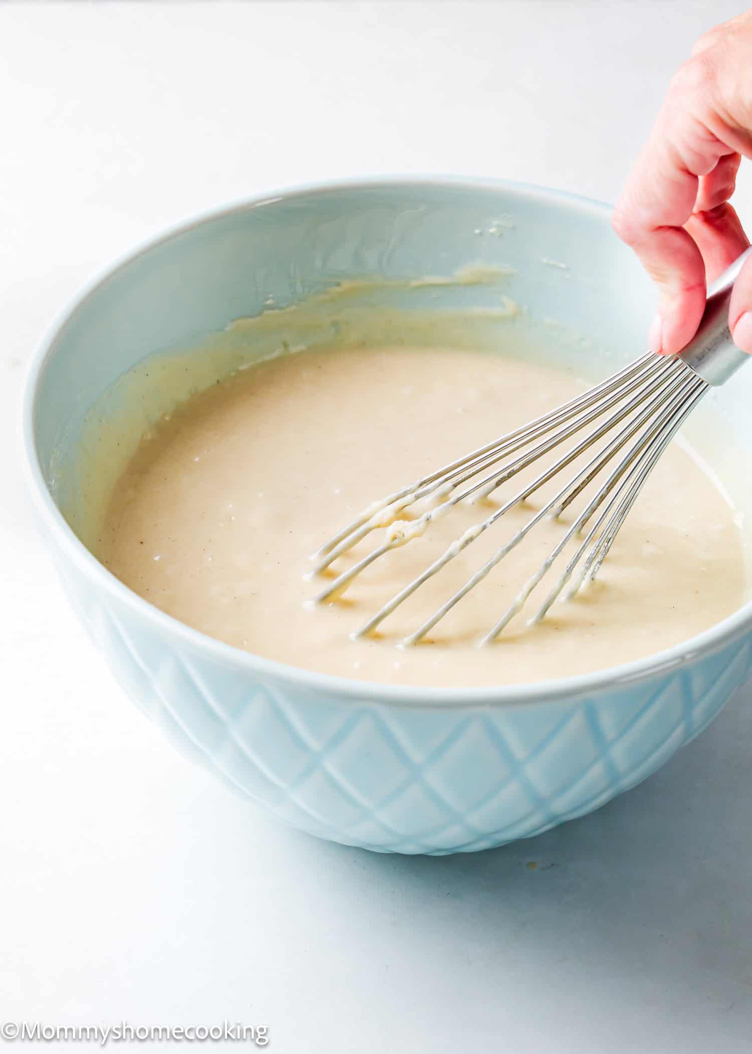 A hand uses a metal whisk to easily mix batter for an egg-free and dairy-free vanilla sheet cake in a light blue bowl on a white surface.