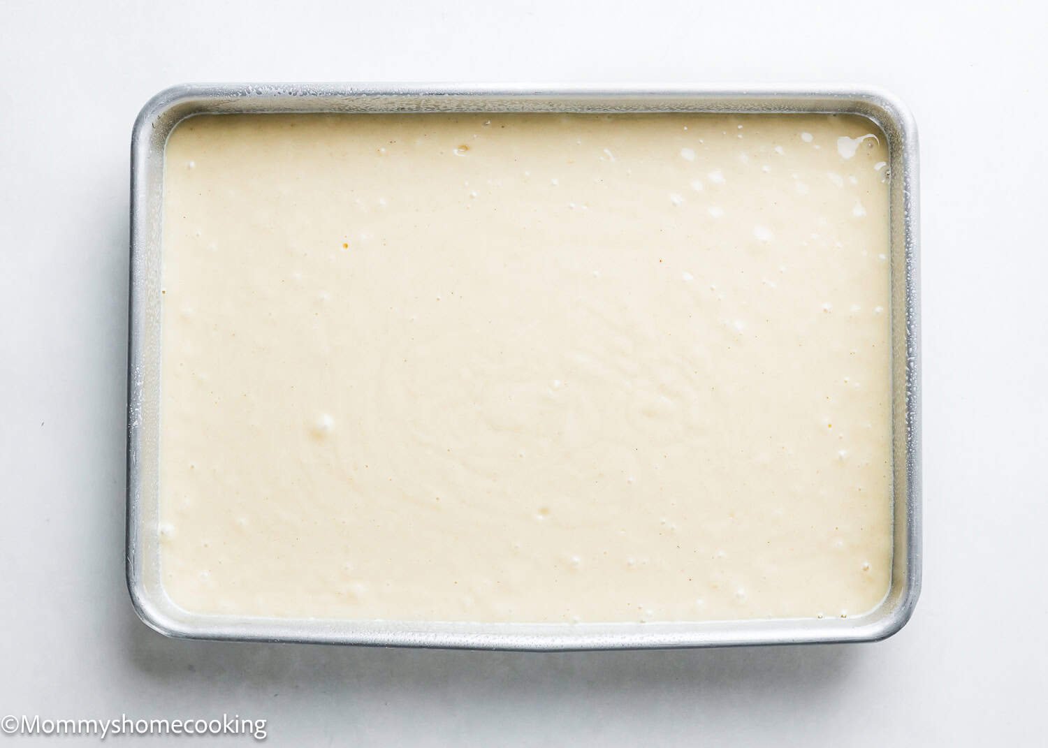 A rectangular baking pan holds a smooth, light beige batter, perfect for an easy, egg-free and dairy-free vanilla sheet cake.