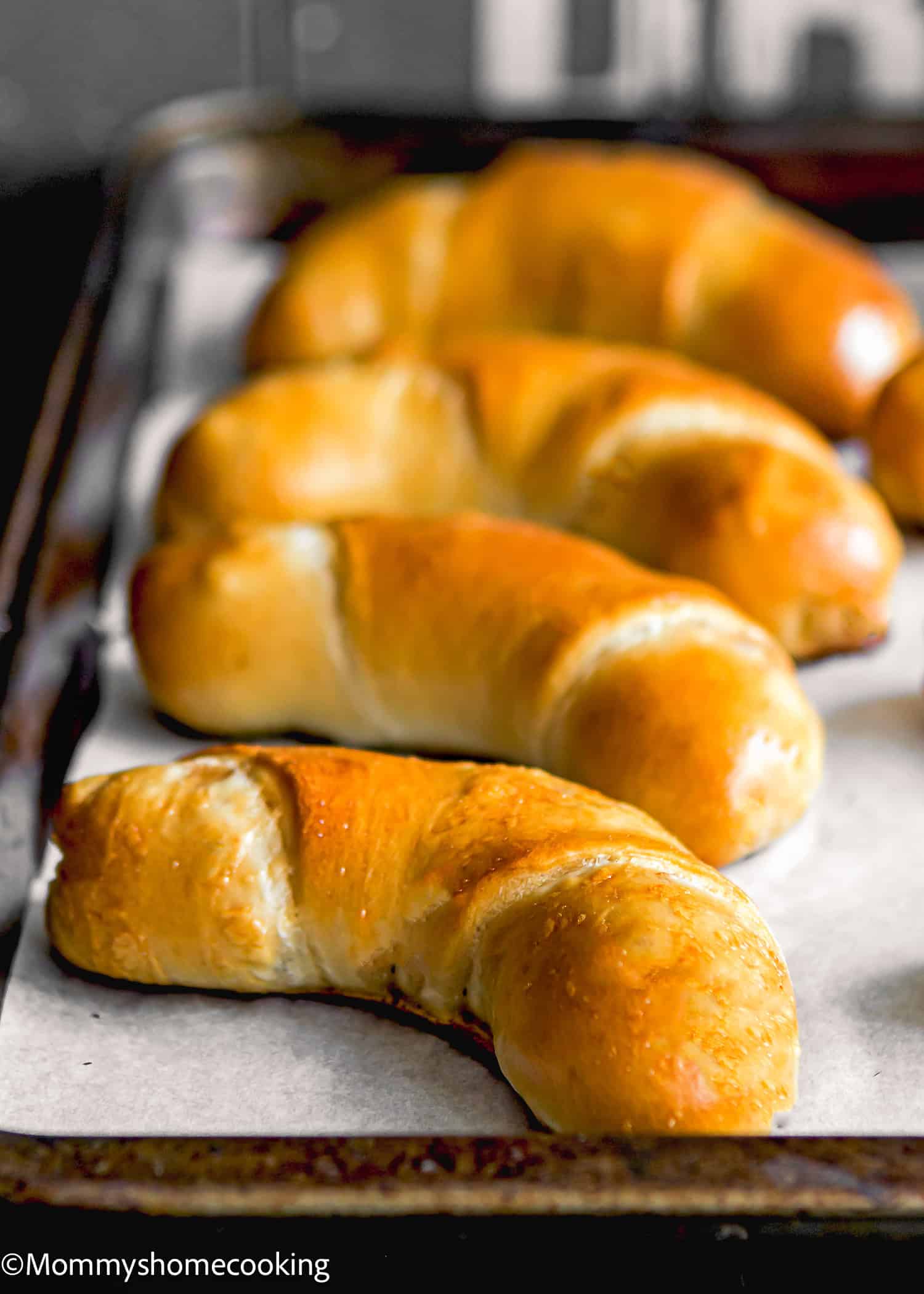 Four freshly baked Venezuelan Cachitos de Jamon rest on a parchment-lined baking tray, their golden crusts hinting at the savory ham filling within.