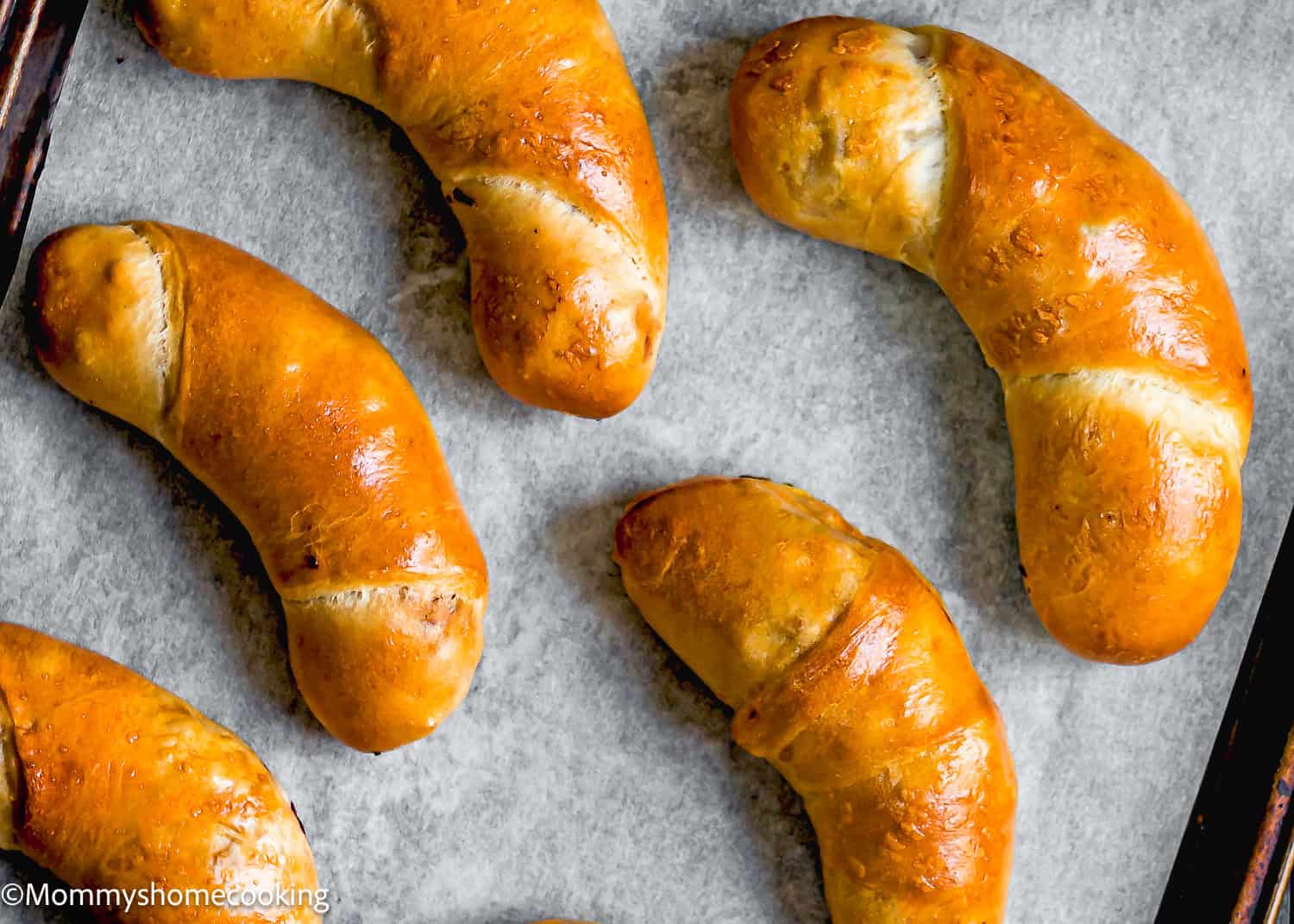 Six freshly baked golden brown croissants, reminiscent of Venezuelan Cachitos de Jamon, rest on a parchment-lined baking sheet.