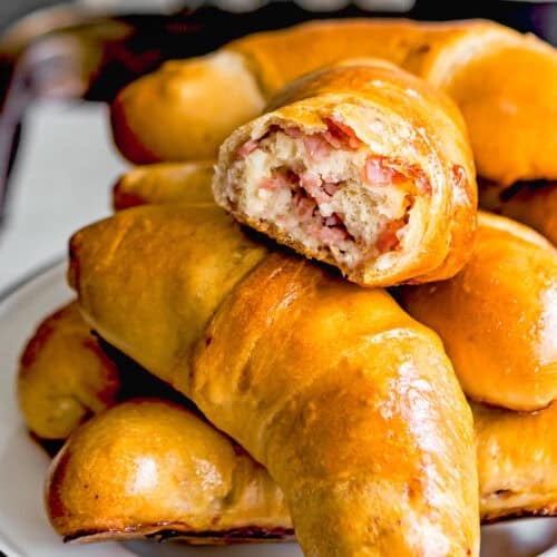 A plate of golden-brown crescent pastries, resembling Venezuelan Cachitos de Jamon, with one on top revealing a savory filling of chopped meat and cheese.