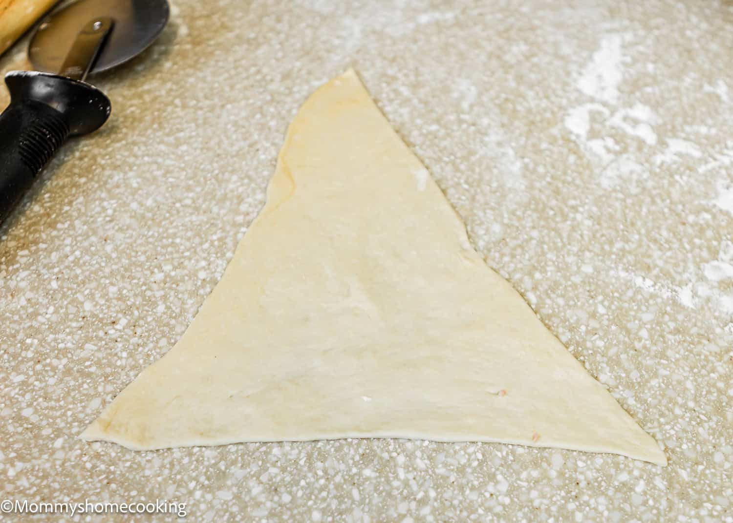 A triangle-shaped dough piece rests on a floured surface, ready to be transformed into delicious Venezuelan Cachitos de Jamon, with a pizza cutter eagerly waiting by its side.
