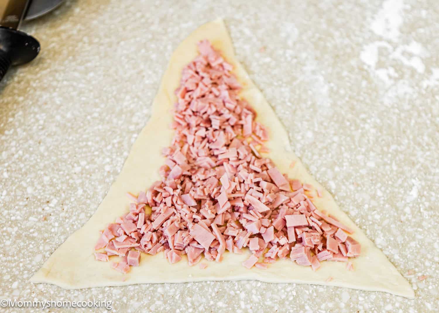 A triangle-shaped Venezuelan Cachitos de Jamon dough with chopped ham filling spread across its surface on a speckled countertop.