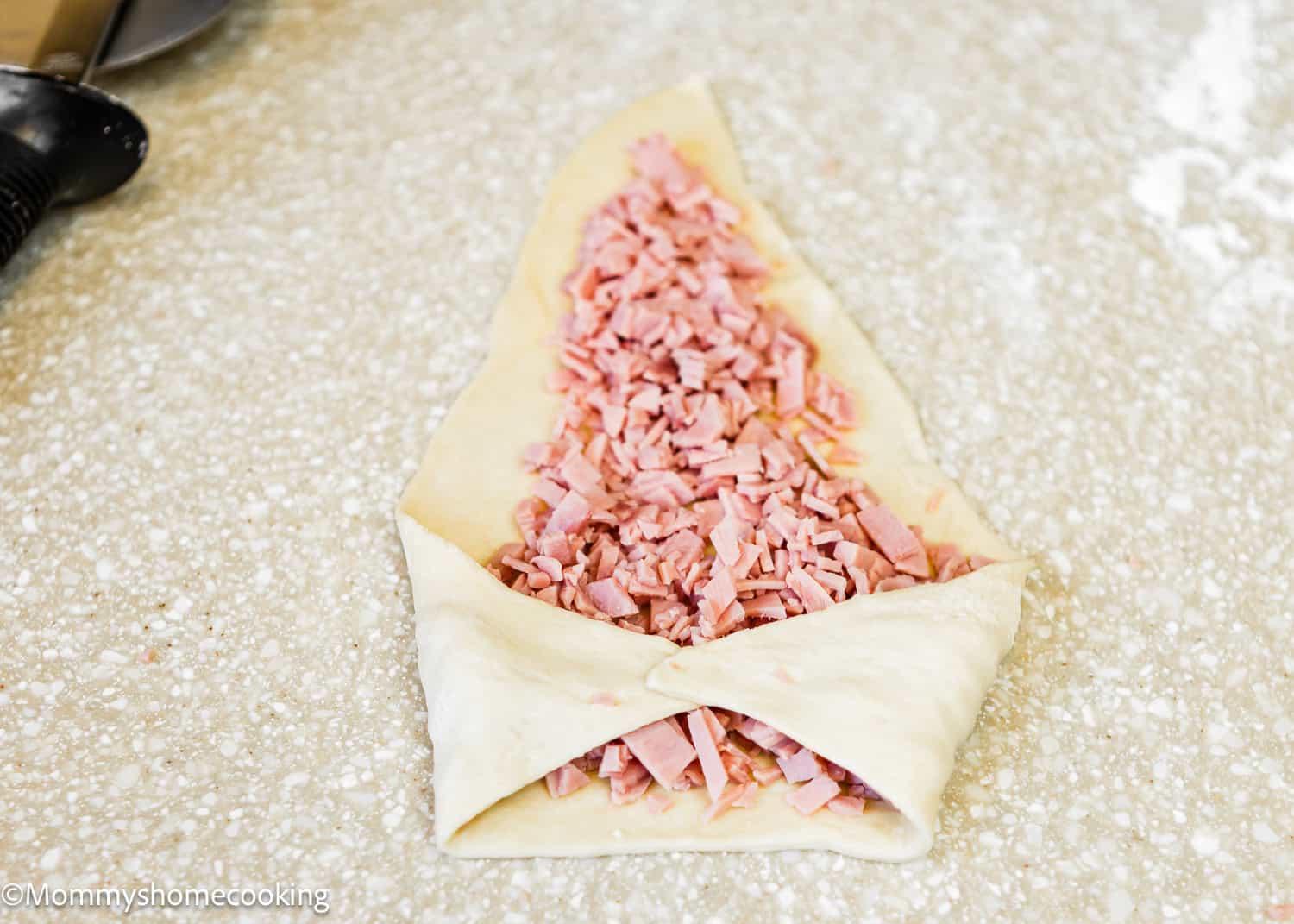 On a speckled countertop sits a triangle-shaped dough, reminiscent of Venezuelan Cachitos de Jamon, filled with chopped ham and edges artfully folded over.