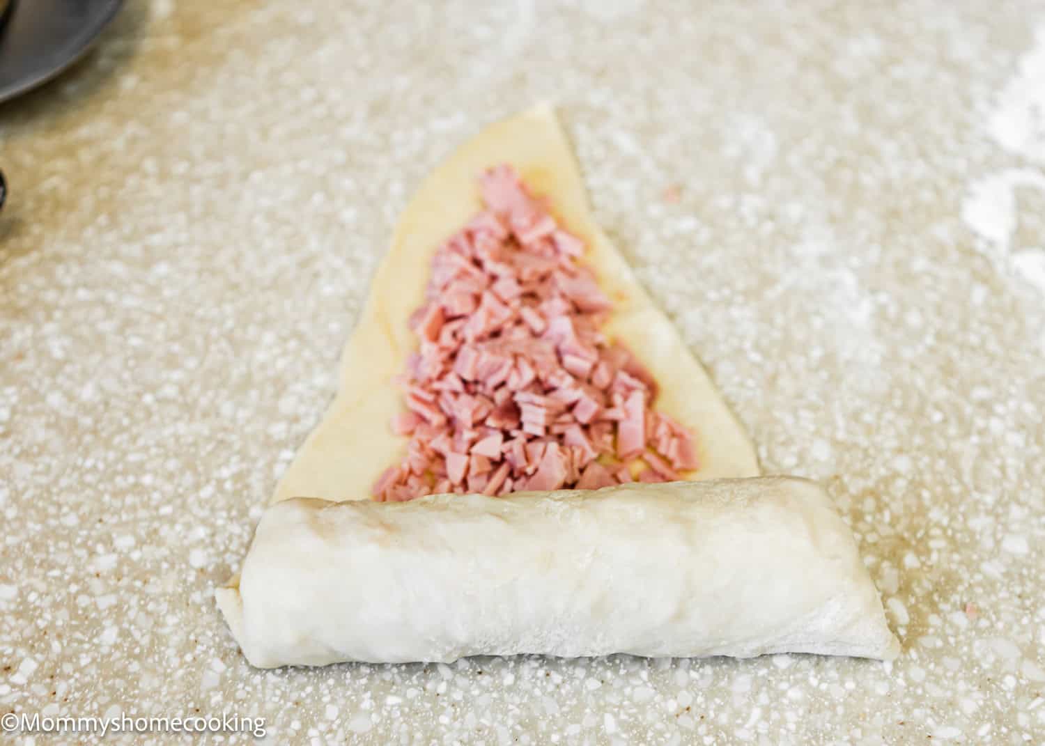 Partially rolled dough with diced ham nestled inside rests on a speckled countertop, reminiscent of the classic Venezuelan Cachitos de Jamon.
