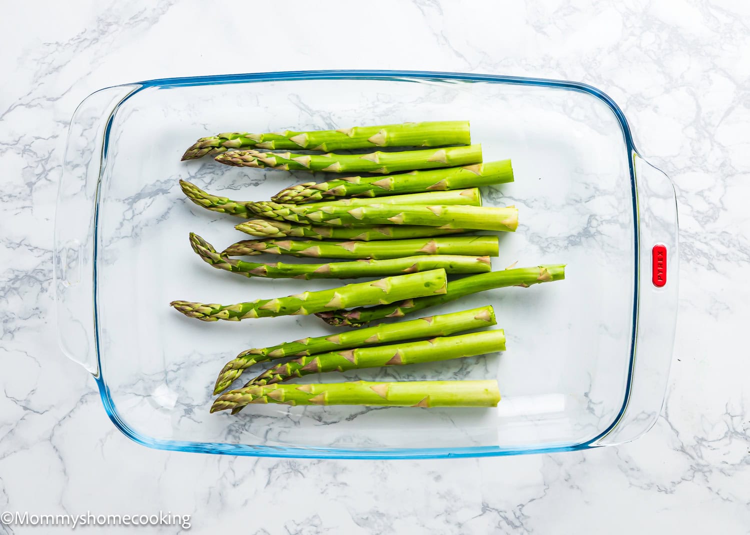 Perfect Air Fryer Asparagus spears are beautifully arranged in a rectangular glass baking dish, resting on a pristine white marble surface.