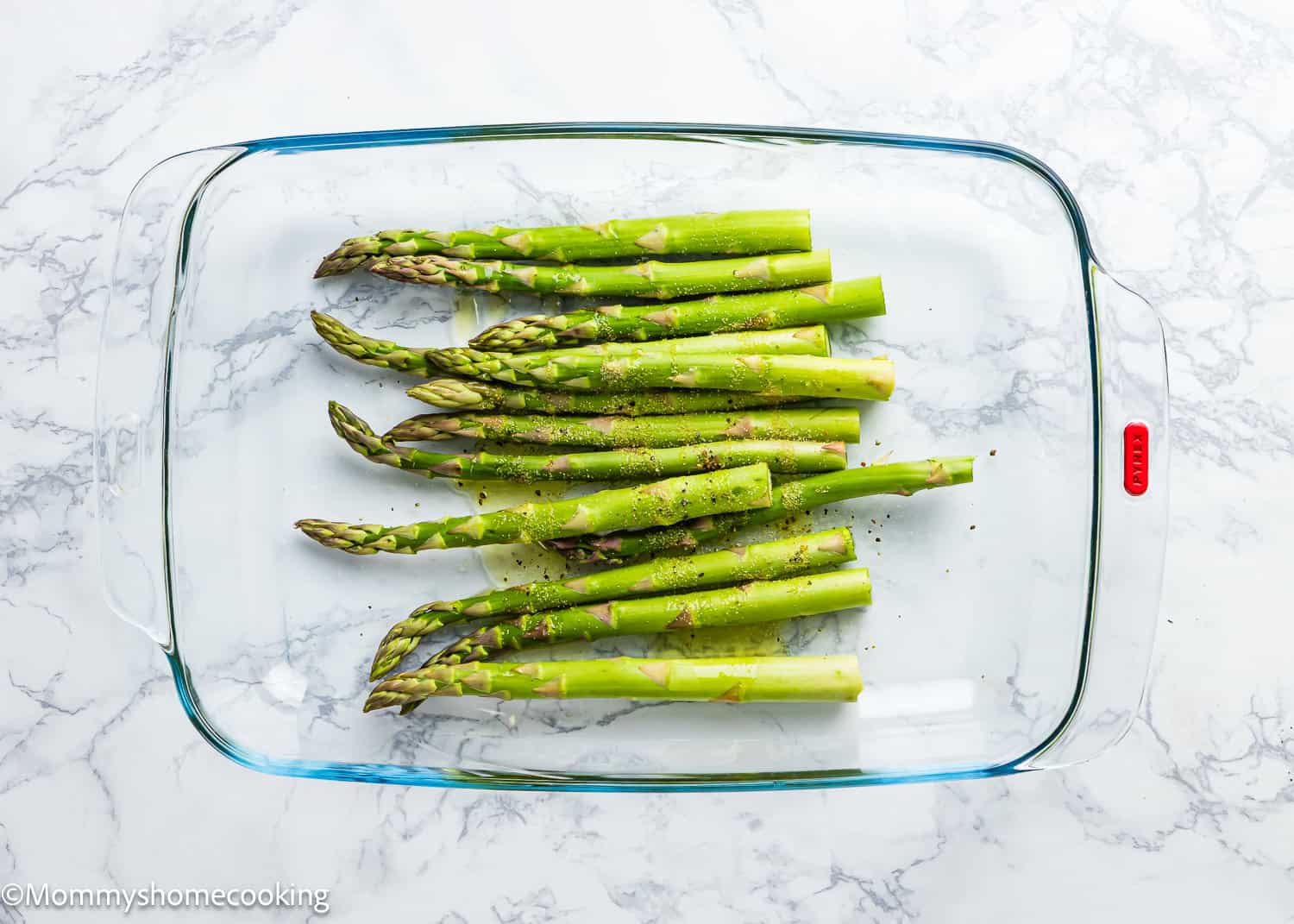 Glass baking dish with fresh asparagus spears, seasoned to perfection with pepper, on a marble surface—perfect air fryer asparagus in the making.