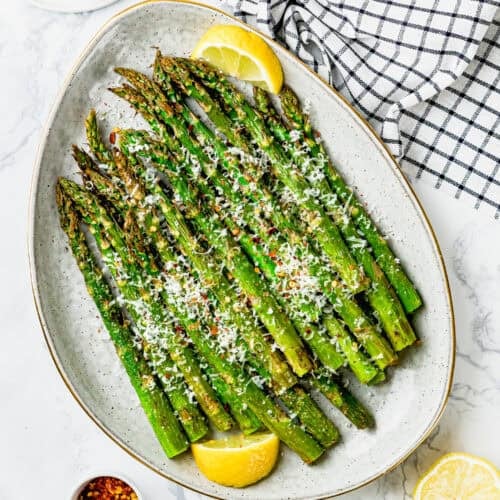 A perfect air fryer asparagus dish awaits with roasted spears topped with grated cheese and seasonings, garnished with lemon wedges. Nearby, a bowl of chili flakes and a block of cheese sit on a checkered cloth, ready to add that extra touch.