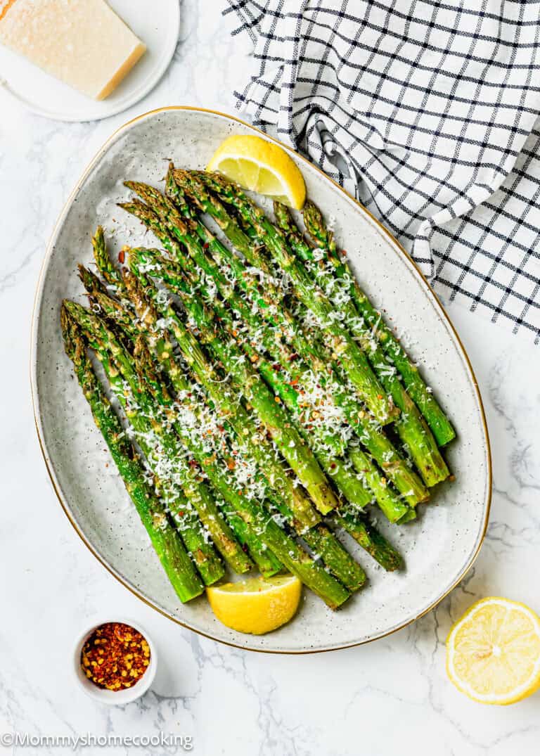 A perfect air fryer asparagus dish awaits with roasted spears topped with grated cheese and seasonings, garnished with lemon wedges. Nearby, a bowl of chili flakes and a block of cheese sit on a checkered cloth, ready to add that extra touch.