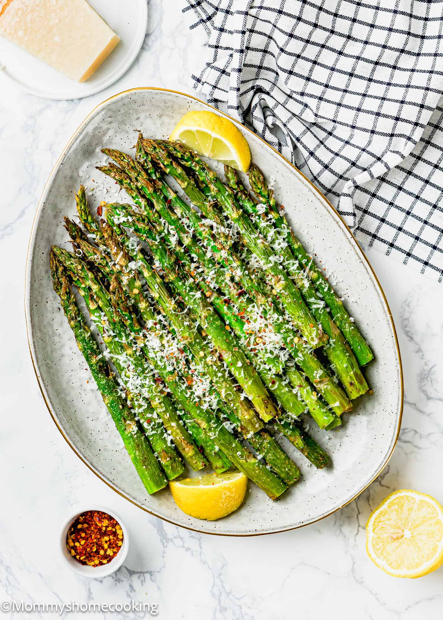 A perfect air fryer asparagus dish awaits with roasted spears topped with grated cheese and seasonings, garnished with lemon wedges. Nearby, a bowl of chili flakes and a block of cheese sit on a checkered cloth, ready to add that extra touch.