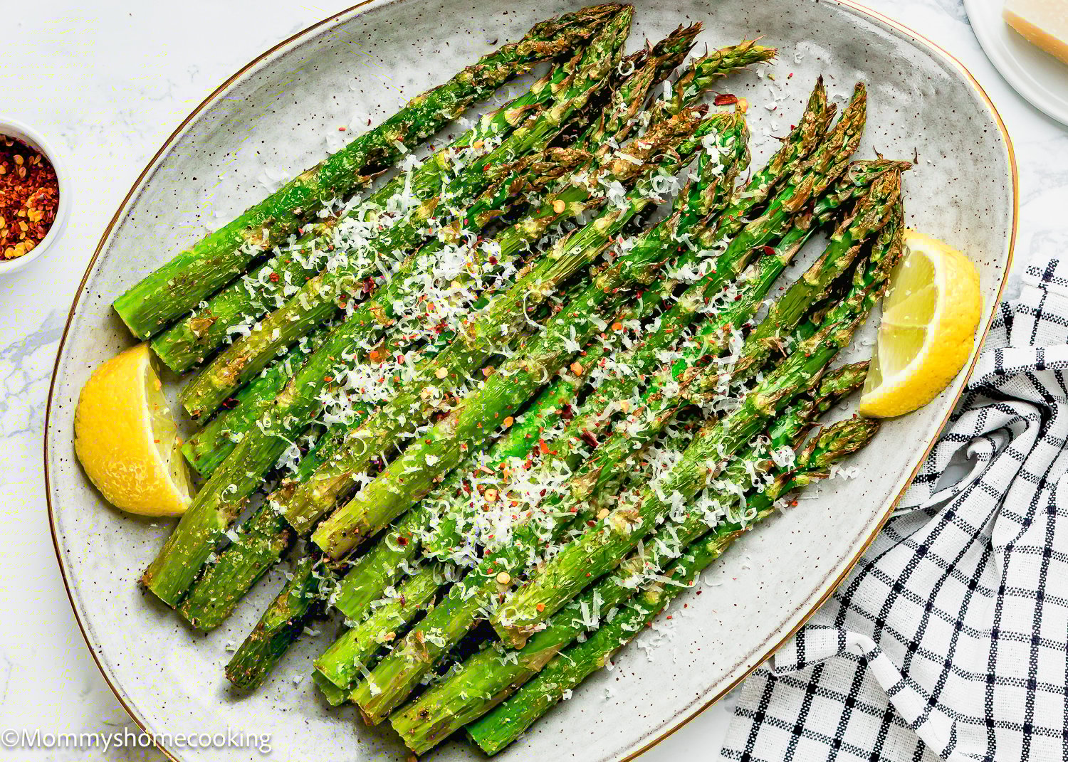 A plate of perfectly air-fried asparagus is topped with grated cheese and red pepper flakes, garnished with lemon wedges. A small bowl of extra red pepper flakes and a checkered cloth sit nearby, ready to accompany this delightful dish.