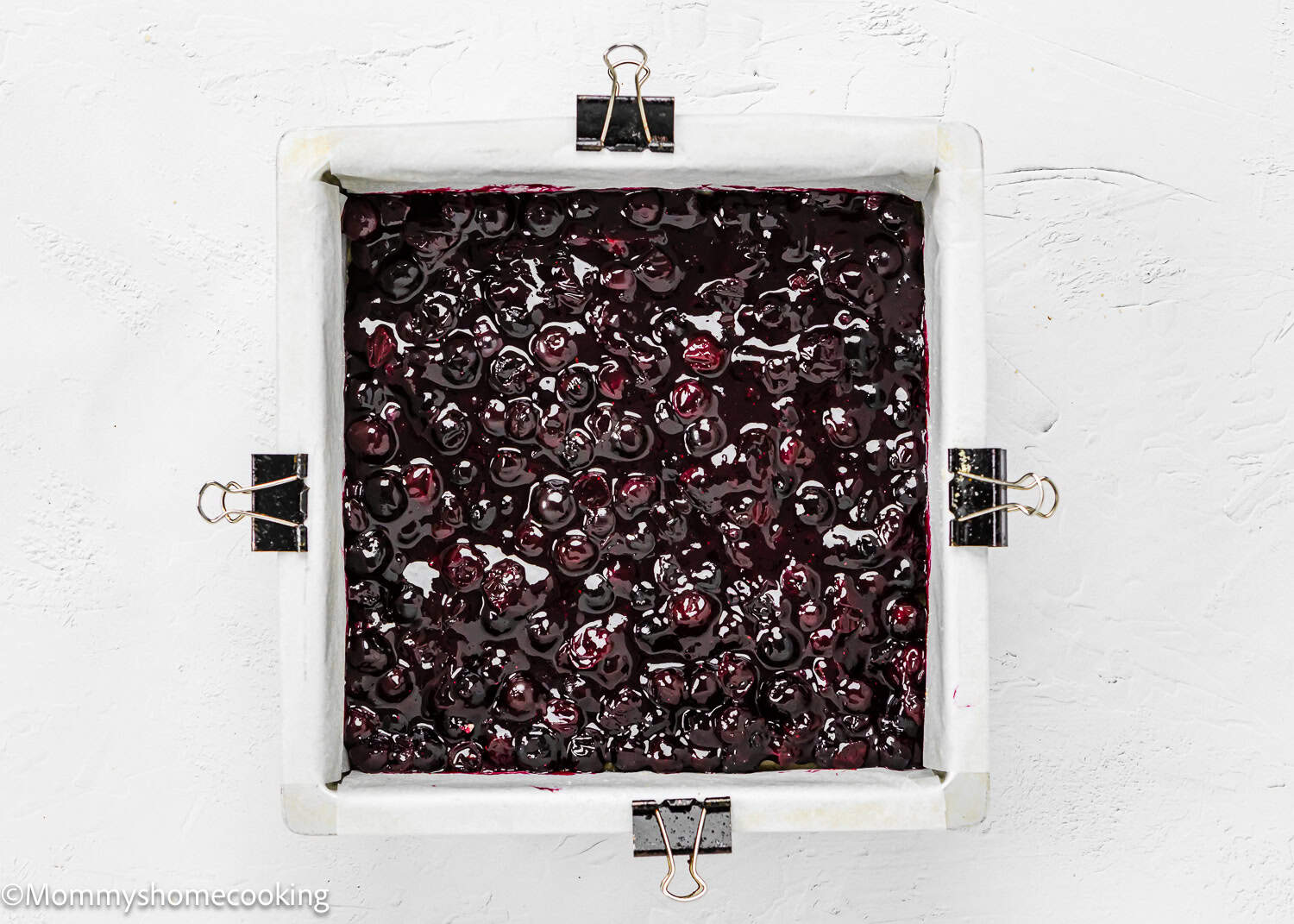 A square baking pan filled with a delicious dark berry mixture, lined with parchment paper secured by binder clips, awaits on a white surface. These Easy Eggless Blueberry Oat Crumb Bars are perfect for any occasion.