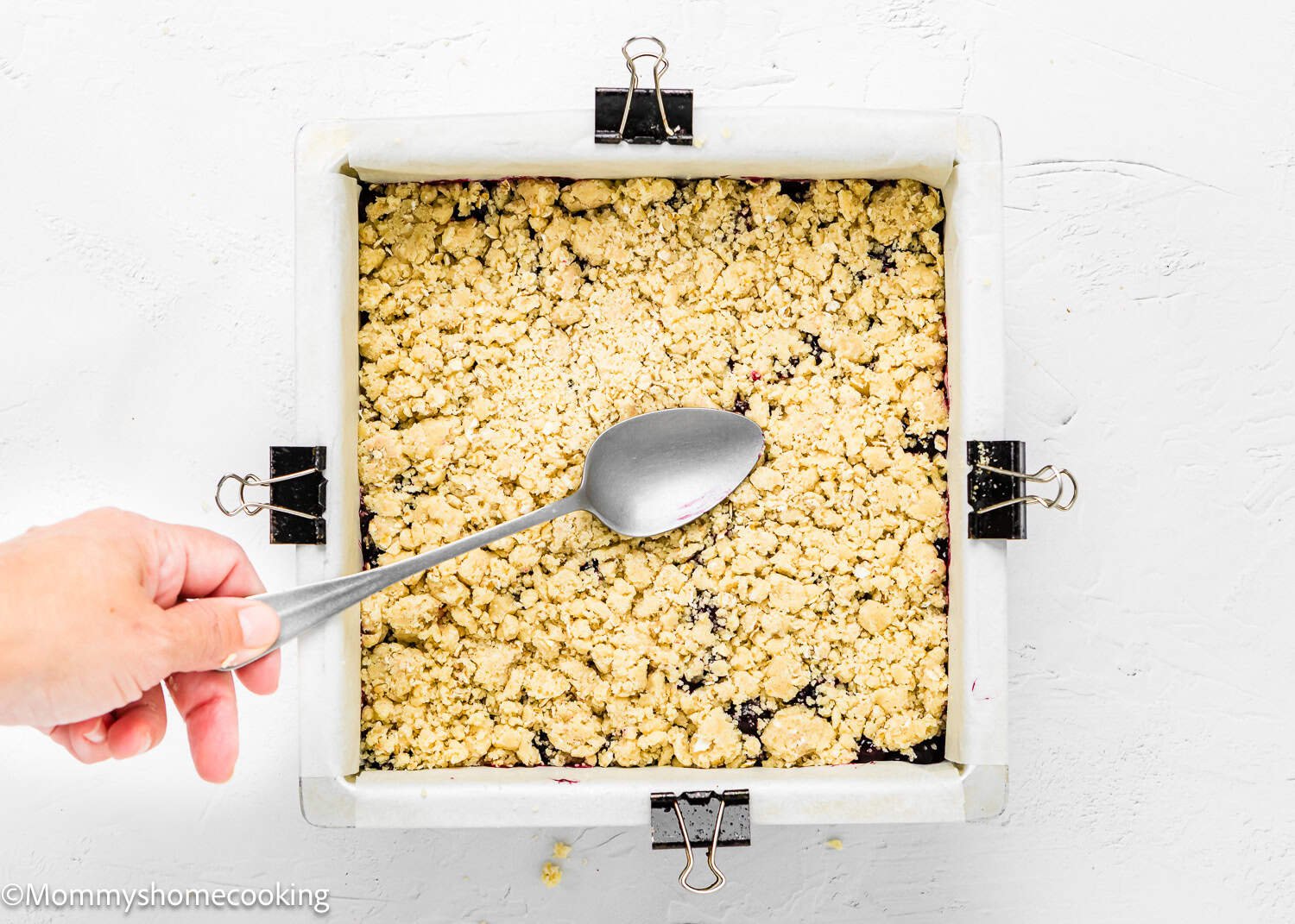 A hand holding a spoon over an unbaked crumble mixture in a square pan prepares the base for easy, eggless blueberry oat crumb bars, secured with clips.
