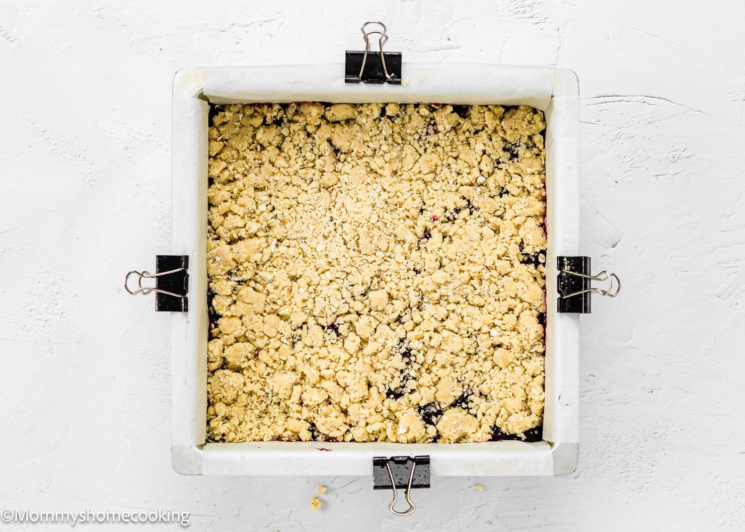 The square baking pan is lined with parchment paper, filled with Easy Eggless Blueberry Oat Crumb Bars, and secured with binder clips on the sides.