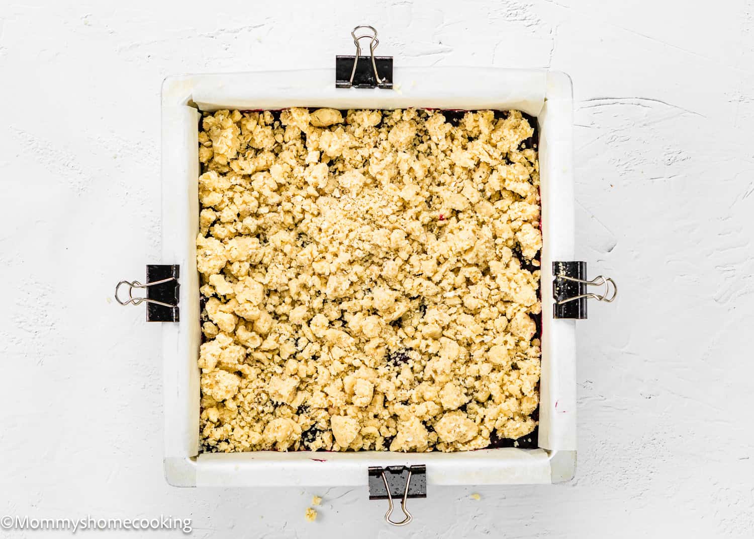 Square baking pan filled with unbaked Easy Eggless Blueberry Oat Crumb Bars, lined with parchment paper and secured with binder clips, on a white surface.
