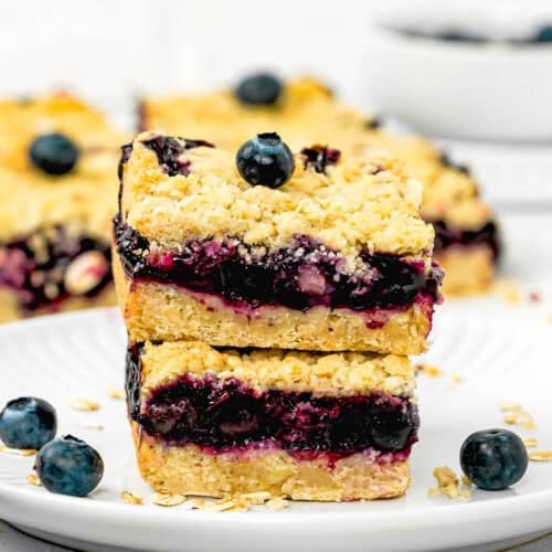 Two easy, eggless blueberry oat crumb bars stacked on a white plate, with oats and fresh blueberries scattered around.