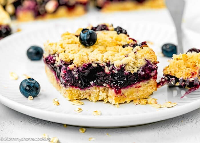 An easy eggless blueberry oat crumb bar sits on a white plate, crowned with a single blueberry. In the background, another bar awaits, while oats are scattered around, adding a rustic touch.